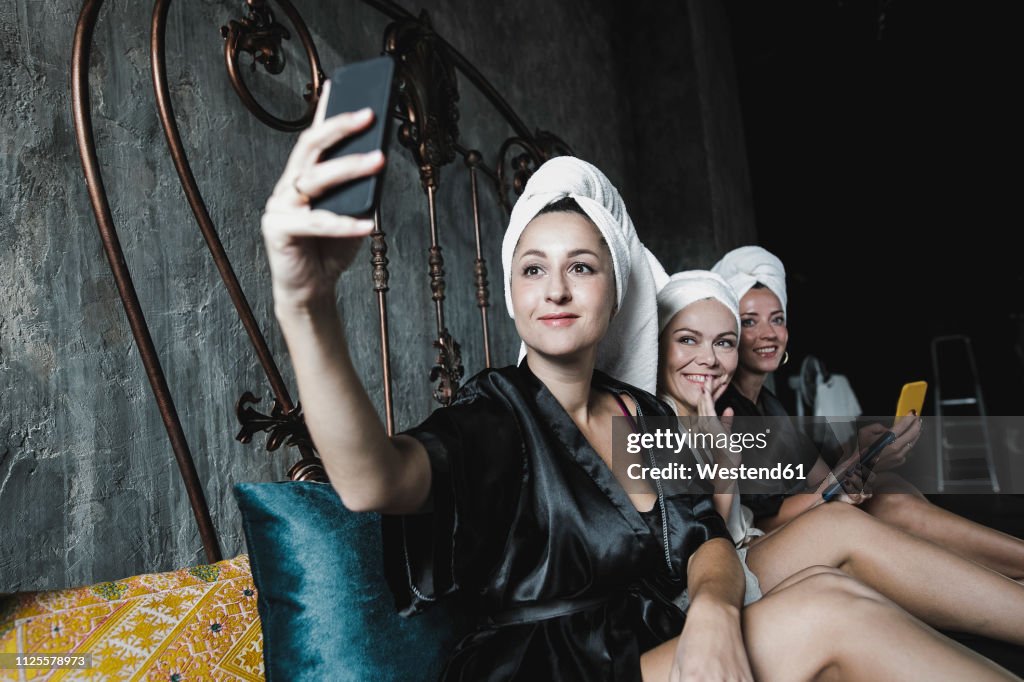 Three women with towels around her heads on bed taking a selfie