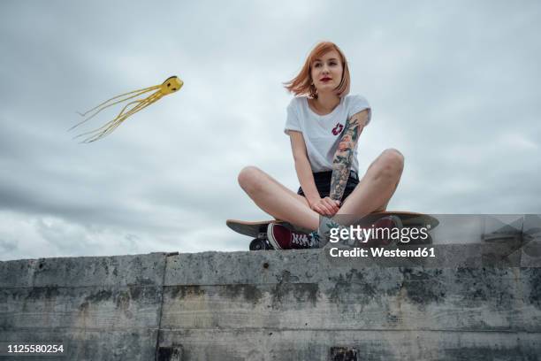 young woman sitting on a concrete wall on carver skateboard with kite in background - stocking tops stock-fotos und bilder