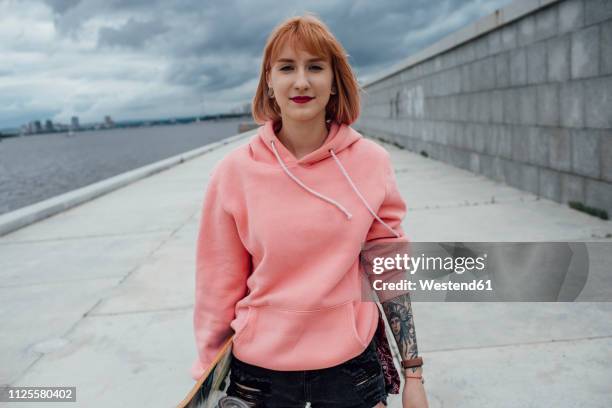 portrait of young woman holding skateboard at the riverside - kapuzenoberteil stock-fotos und bilder