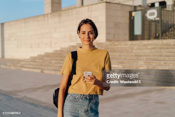 portrait of smiling young woman holding cell phone outdoors - young women stock pictures, royalty-free photos & images