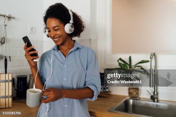 woman with headphones, using smartphone and drinking coffee for breakfast in her kitchen - woman drinking phone kitchen stockfoto's en -beelden