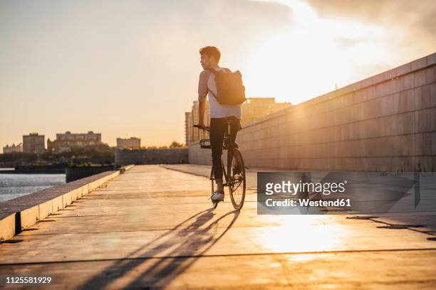 young man with backpack riding bike on waterfront promenade at the riverside at sunset - city bike foto e immagini stock