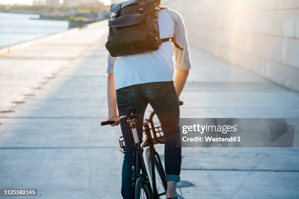 rear view of young man with backpack riding bike on waterfront promenade at the riverside at sunset - concrete wall stock pictures, royalty-free photos & images