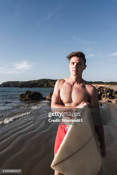 france, brittany, young man carrying surfboard standing at the sea - swimming shorts stock pictures, royalty-free photos & images