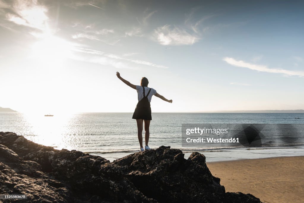 France, Brittany, rear view of young woman standing on rock at the beach at sunset