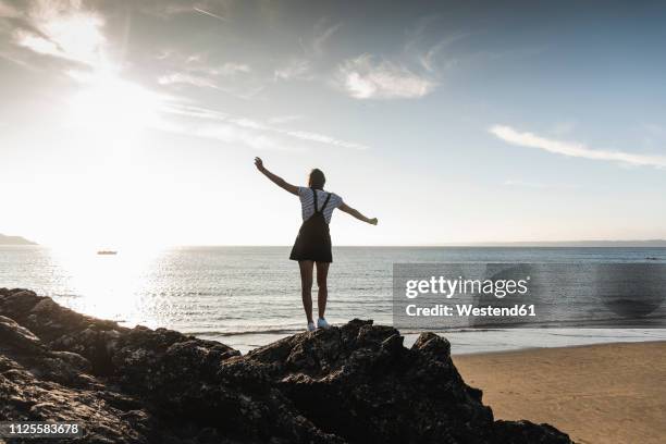 france, brittany, rear view of young woman standing on rock at the beach at sunset - arms outstretched silhouette stock pictures, royalty-free photos & images