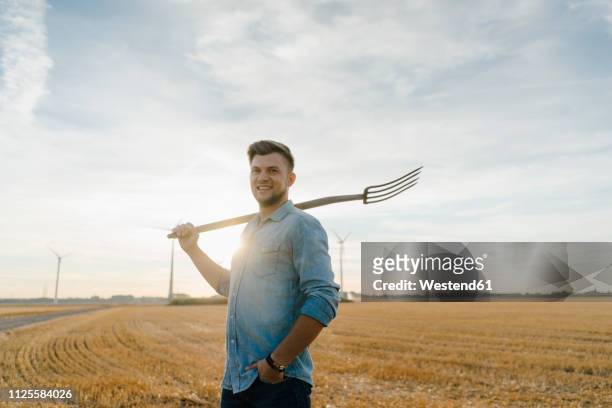 portrait of smiling young man holding pitchfork standing on stubble field - pitchfork agricultural equipment bildbanksfoton och bilder