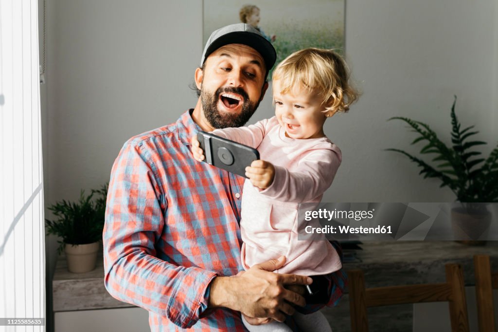 Father holding his little daughter on his arm, playing with his smartphone
