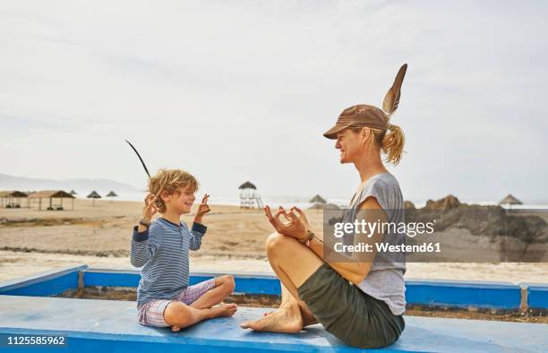 chile, arica, happy mother in yoga pose sitting with son on wall on the beach - arica fotografías e imágenes de stock