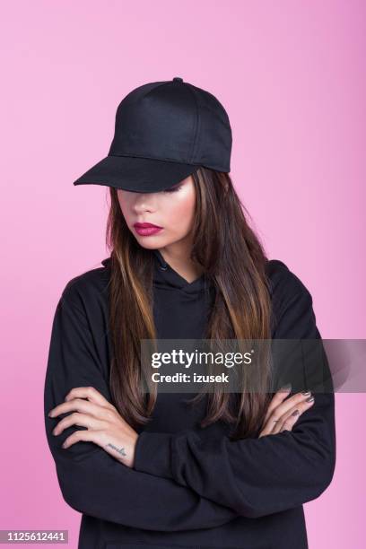 mujer joven en camisa con capucha negra y tapa contra fondo rosa - gorra fotografías e imágenes de stock