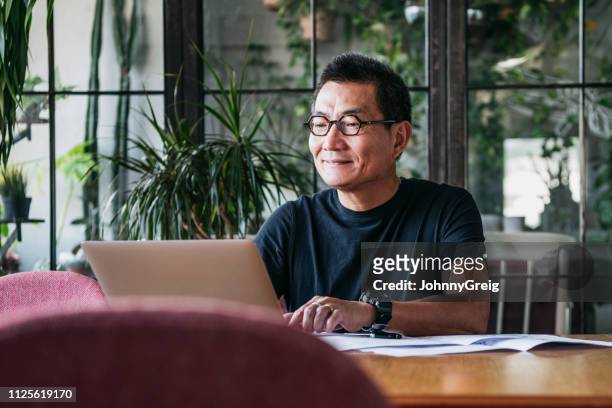 lachende chinese man aan het werk op laptop thuis - mature men stockfoto's en -beelden
