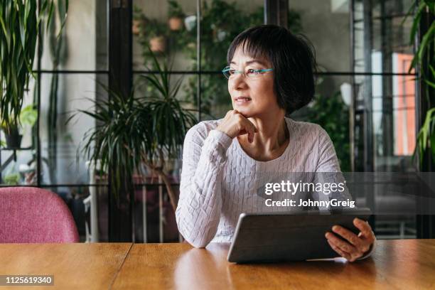 chinese woman in her 60s using tablet with hand on chin - brood stock pictures, royalty-free photos & images