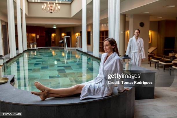 smiling young woman in bathrobe sitting at the poolside in a spa with man in background - accappatoio foto e immagini stock