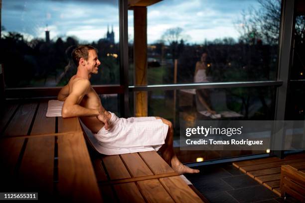 man sitting in a sauna with woman reflected in panoramic window - sauna stock-fotos und bilder