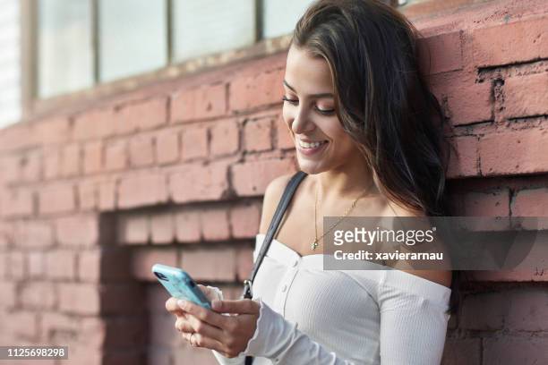 mulher sorridente usando telefone inteligente contra a parede de tijolos - off shoulder - fotografias e filmes do acervo