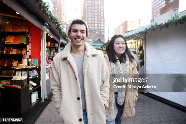 two millennials, one caucasian male and one asian female, enjoy their walk through an outdoor holiday market in the city. - long coat stock pictures, royalty-free photos & images