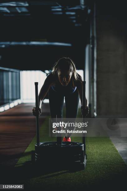 woman doing sled training in a dark gym - sled push stock pictures, royalty-free photos & images