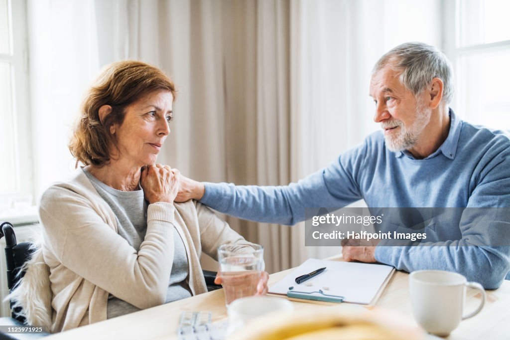 A sad senior couple in wheelchair sitting at the table at home, talking.