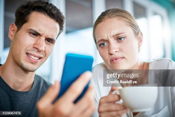 young couple frowning at mobile phone in coffee shop - offense stock pictures, royalty-free photos & images