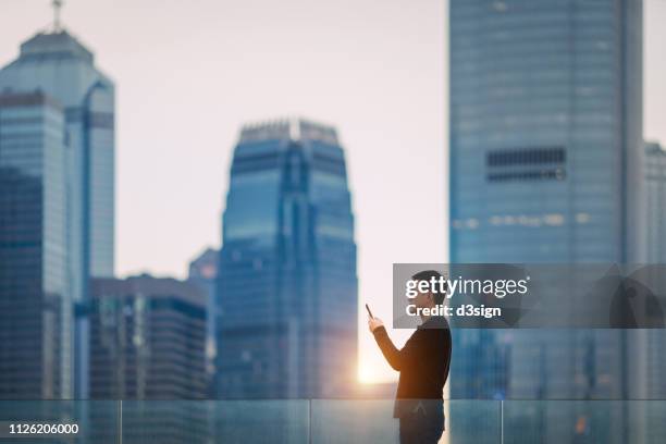 confident young asian businessman using smartphone against hong kong cityscape with contemporary skyscrapers in financial district - central asia stock-fotos und bilder