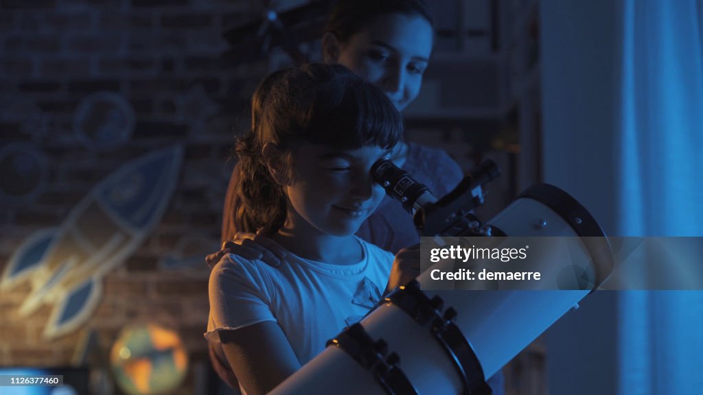Cute sisters watching the stars together at home using a telescope, family and leisure concept