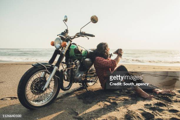 guy sitting by his bike on the beach taking pictures with instant camera - muscle men at beach stock pictures, royalty-free photos & images