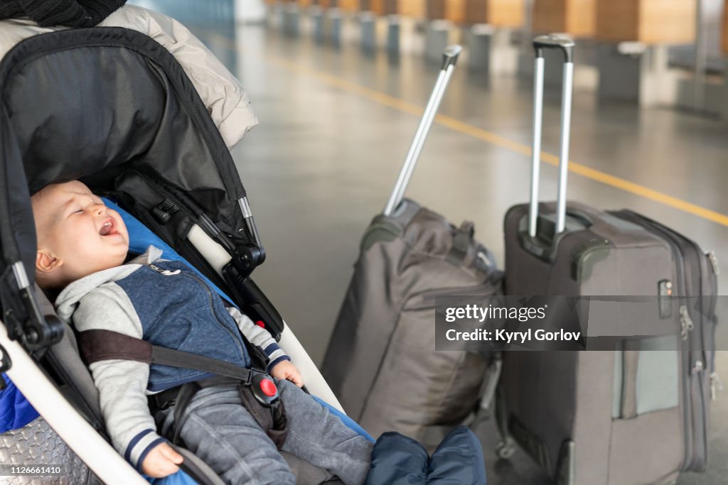 Bambino urlante seduto nel passeggino vicino ai bagagli al terminal dell'aeroporto. Bambino in carrozza vicino al banco del check-in. Bambini lacrime, panico e isterica. Concetto di viaggiare con bambini piccoli