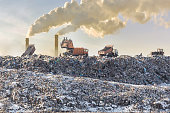 Dump trucks unloading garbage over vast landfill. Smoking industrial stacks on background. Environmental pollution. Outdated method of waste disposal. Survival of times past