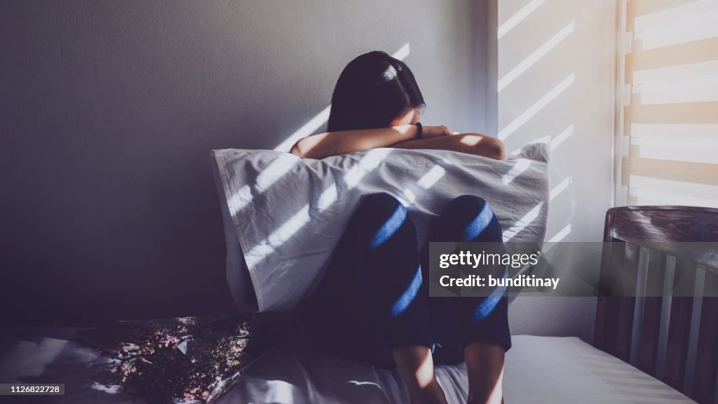 Asian women are sitting hugging their knees in bed. Feeling sad, disappointed in love In the dark bedroom and sunlight from the window through the blinds.Vintage tone.