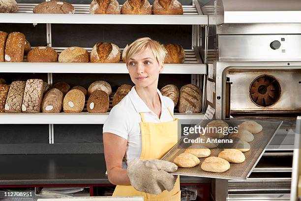 a woman pulling a tray of freshly baked rolls in a bakery - part of a series foto e immagini stock
