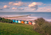 Impressive winter clouds in a cool blue sky over the beach huts and natural spit of land that stretches out to sea on the beach in Tankerton, Whitstable, Kent, UK. A three people are strolling on the natural and locally named  'Street'