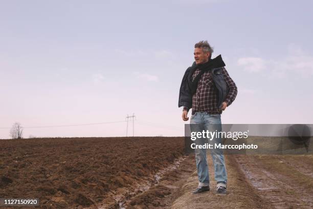 handsome adult farmer standing next to his plowed farm fields on windy day in late winter - air serbia stock pictures, royalty-free photos & images