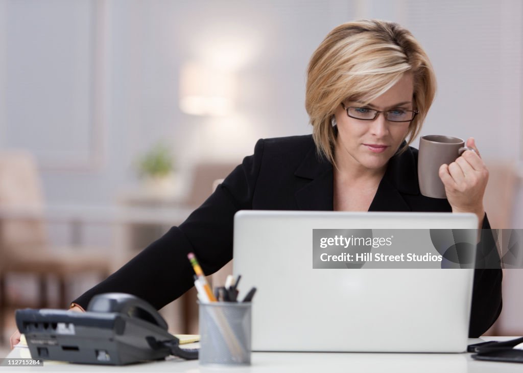 Caucasian businesswoman using laptop in home office and drinking coffee