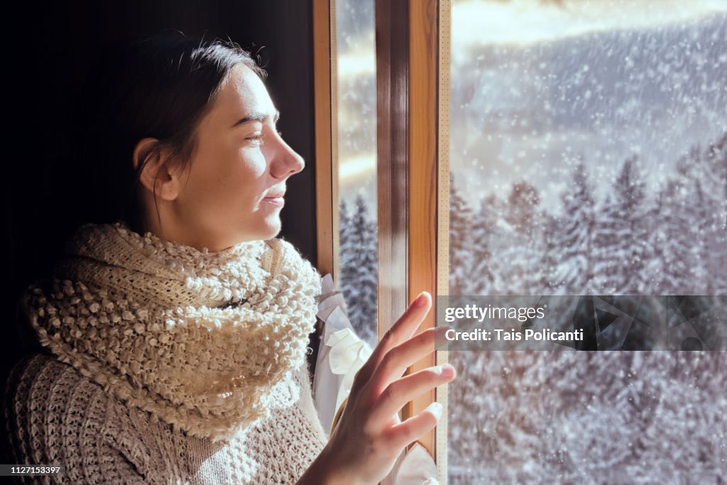 Young woman looking to the forest from her window while it snows - Bavaria, Germany