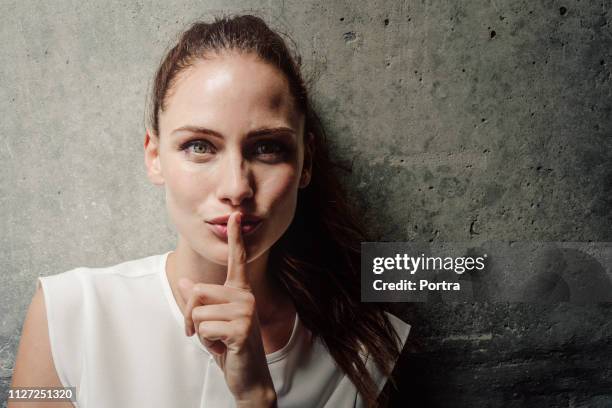 young woman with finger on lips against wall - quiet gesture stock pictures, royalty-free photos & images