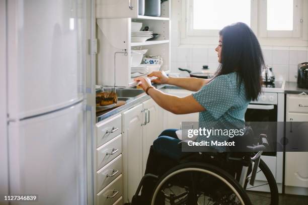 side view of young disabled woman holding cabinet in kitchen at home - rolstoelvriendelijk stockfoto's en -beelden