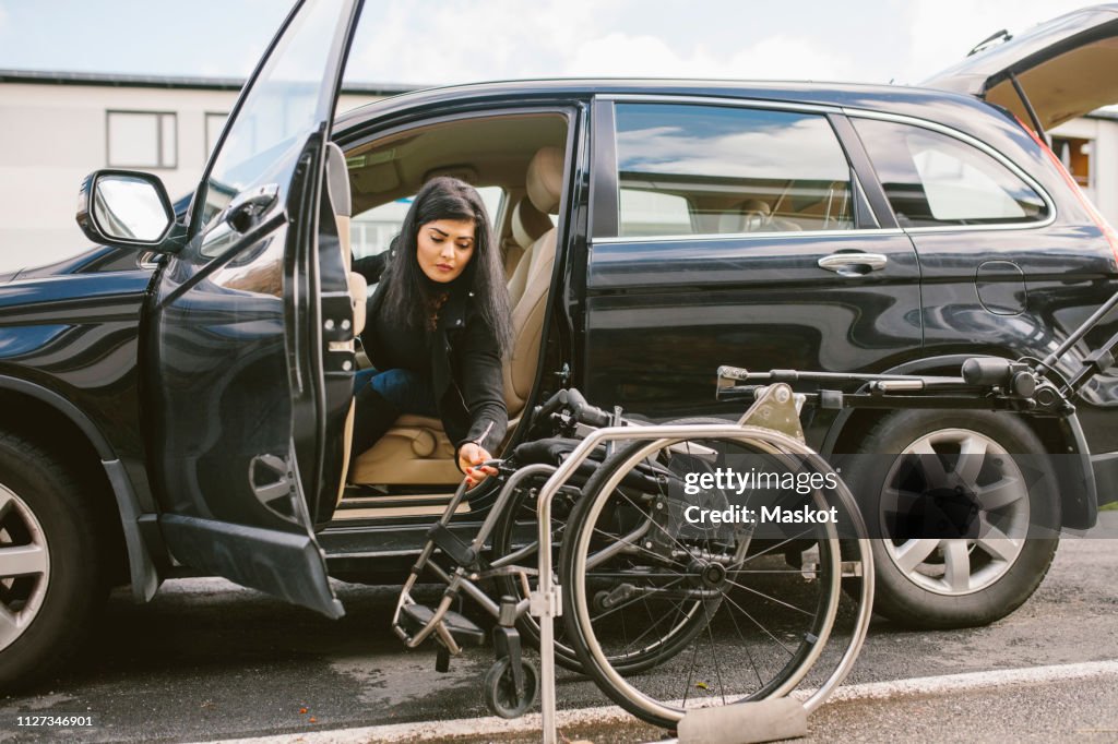 Young disabled woman holding wheelchair by car on roadside in city