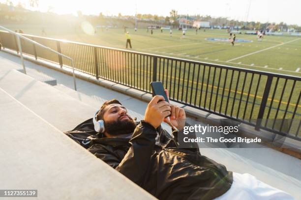 young man lying on bench in stadium using mobile phone - soccer field park stock pictures, royalty-free photos & images