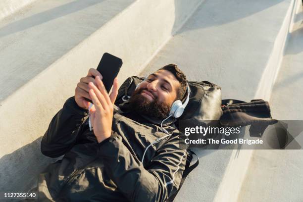 young man lying on bench in stadium listening to music on headphones - young man listening to music on smart phone outdoors stockfoto's en -beelden