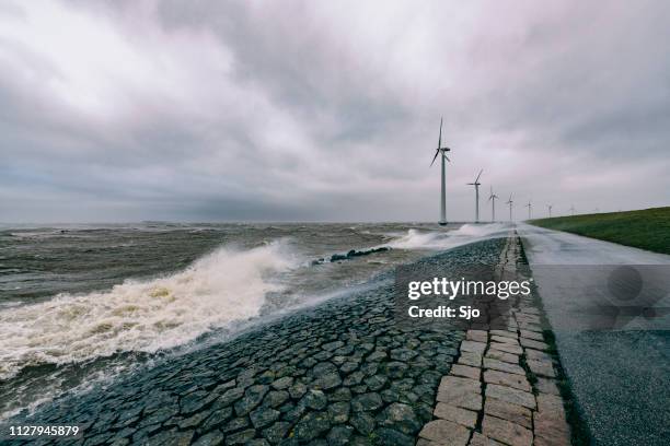 wind turbines on land and offshore in a storm with waves hitting a levee - overcast ocean stock pictures, royalty-free photos & images