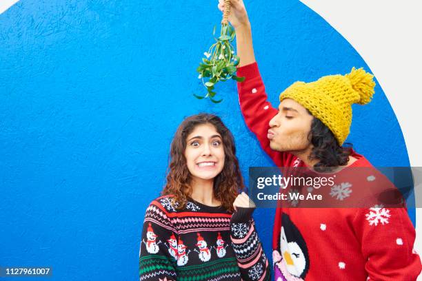 young man holds mistletoe over woman who is making a face - weihnachtspullover stock-fotos und bilder