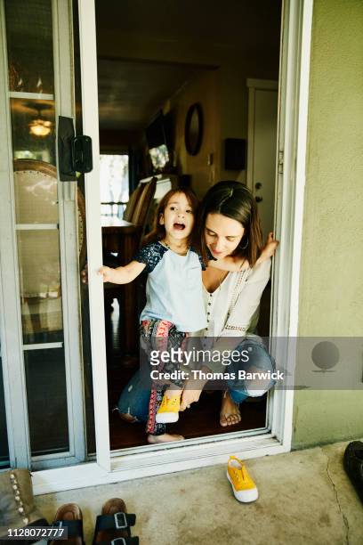 Mother helping young daughter put on shoes while standing at door to back yard
