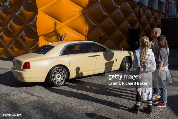 Rolls-Royce limousine drives past less wealthy onlookers opposite the temporary renovation hoarding of luxury brand Louis Vuitton in New Bond Street,...