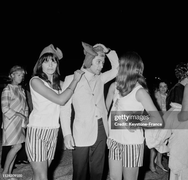 Entre deux charmantes jeunes filles, Marcel Amont se coiffe d'un bonnet phrygien lors d'une fête à Antibes, France, le 11 juillet 1968.