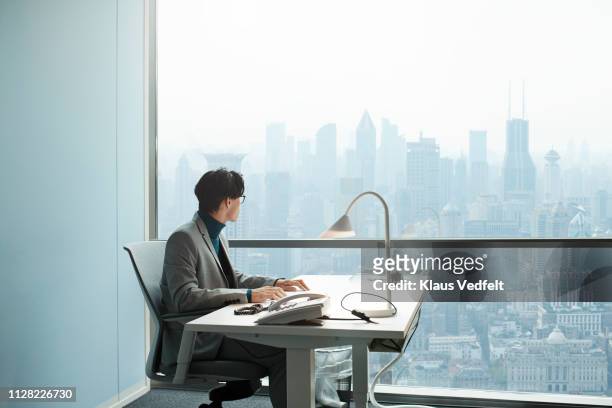 businessman sitting in office and looking out on stunning shanghai skyline - portrait of pensive young businessman wearing glasses stock-fotos und bilder