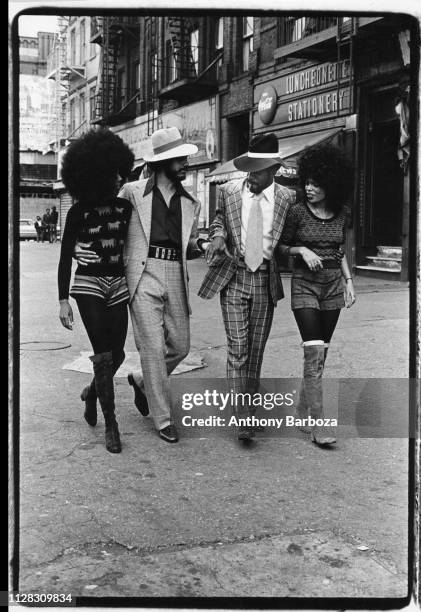 View of four people, two men and two women, as they walk along an unidentified street in Harlem, New York, New York, 1971. The two women both wear...