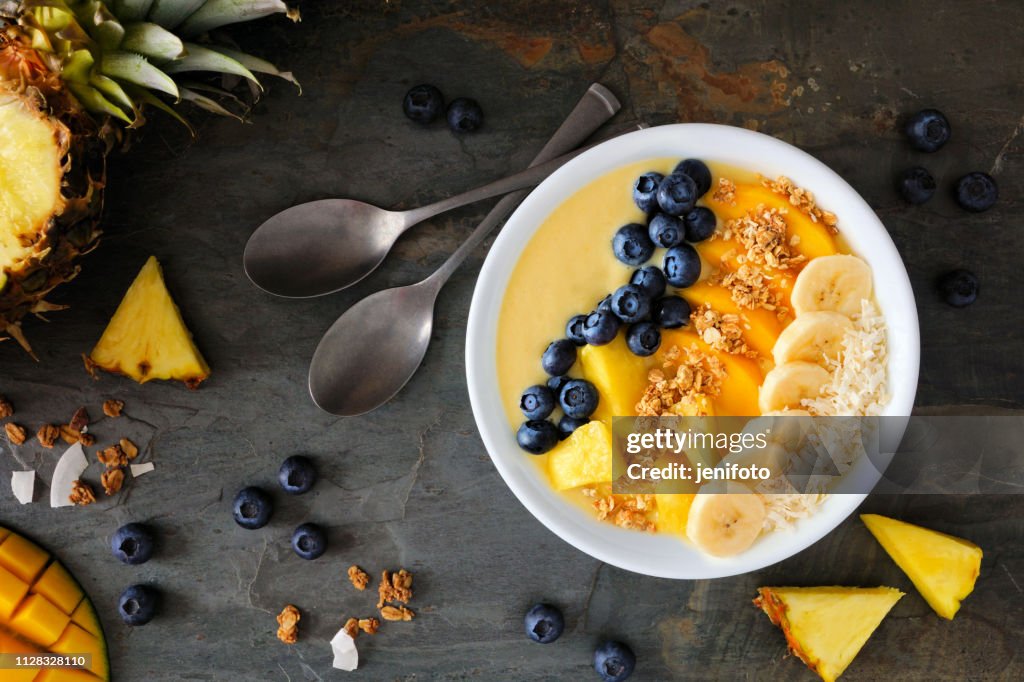 Pineapple, mango smoothie bowl with coconut, bananas, blueberries and granola, top view table scene on slate