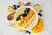Pineapple, mango smoothie bowl with coconut, bananas, blueberries and granola, above view on a bright background
