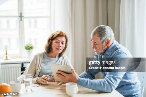 a senior woman in wheelchair sitting at the table at home, taking painkillers and talking to her husband. - mann tabletten stock-fotos und bilder