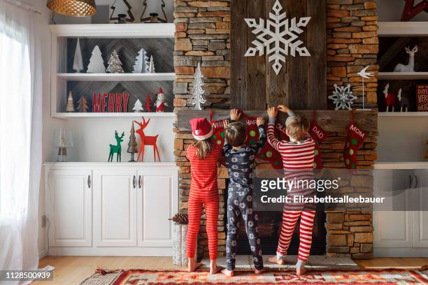 three children hanging up christmas stockings on a fireplace - calza della befana foto e immagini stock